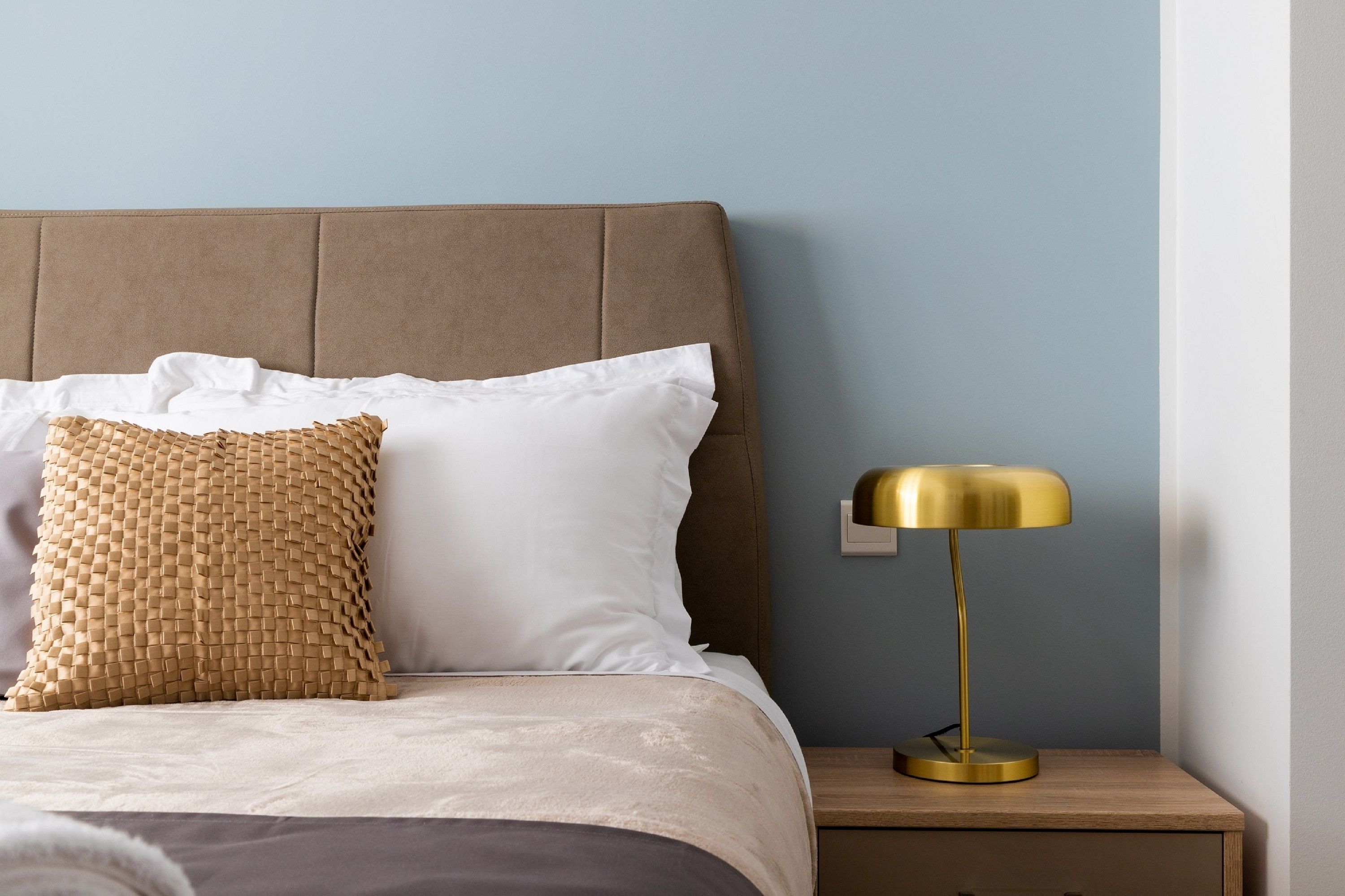 A bed with a brown headboard, white pillows, and a woven throw. A gold lamp is on a wooden nightstand beside the bed.