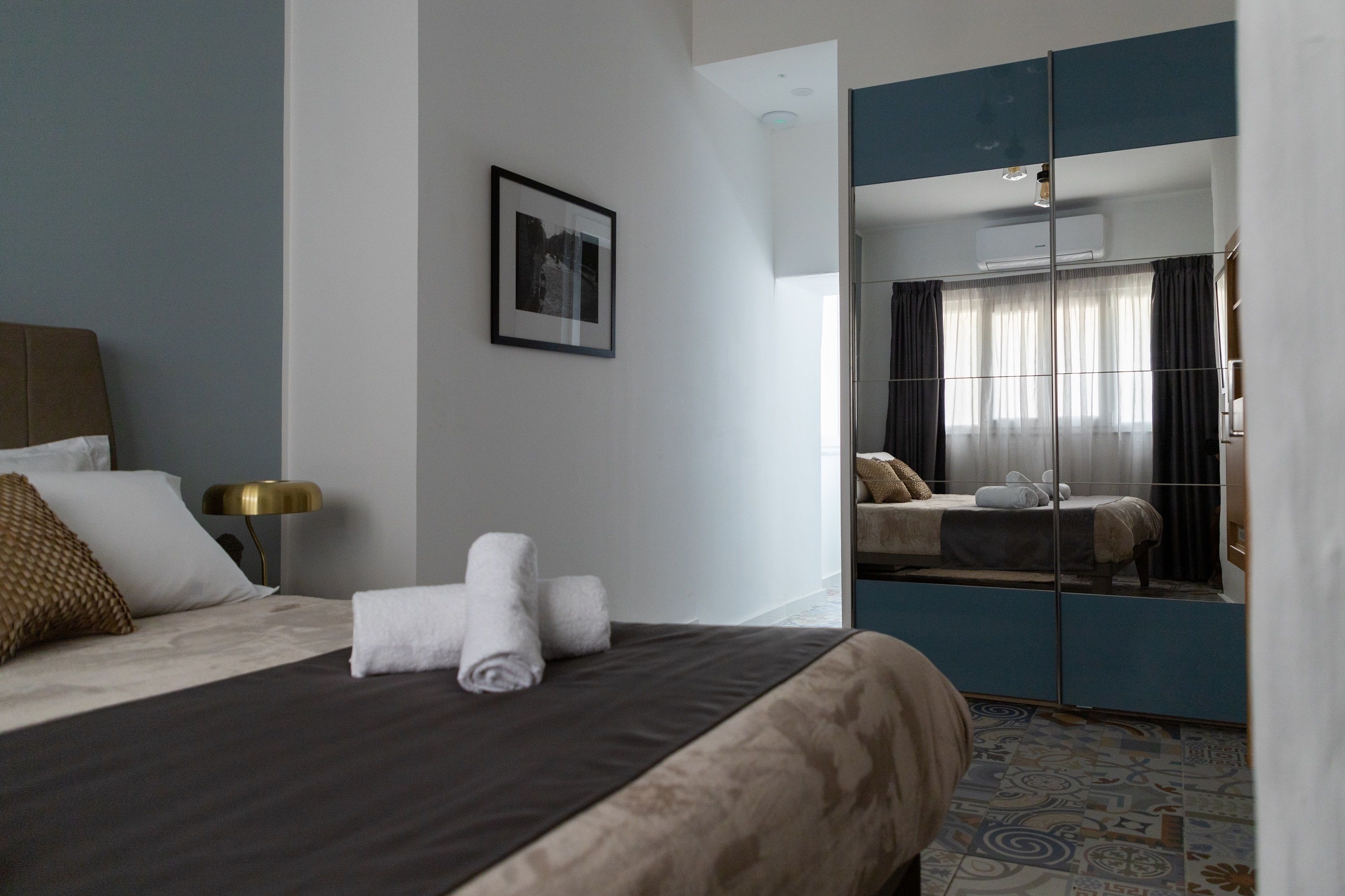 A hotel room with a bed, a bedside lamp, a framed picture on the wall, and a mirror reflecting the room.