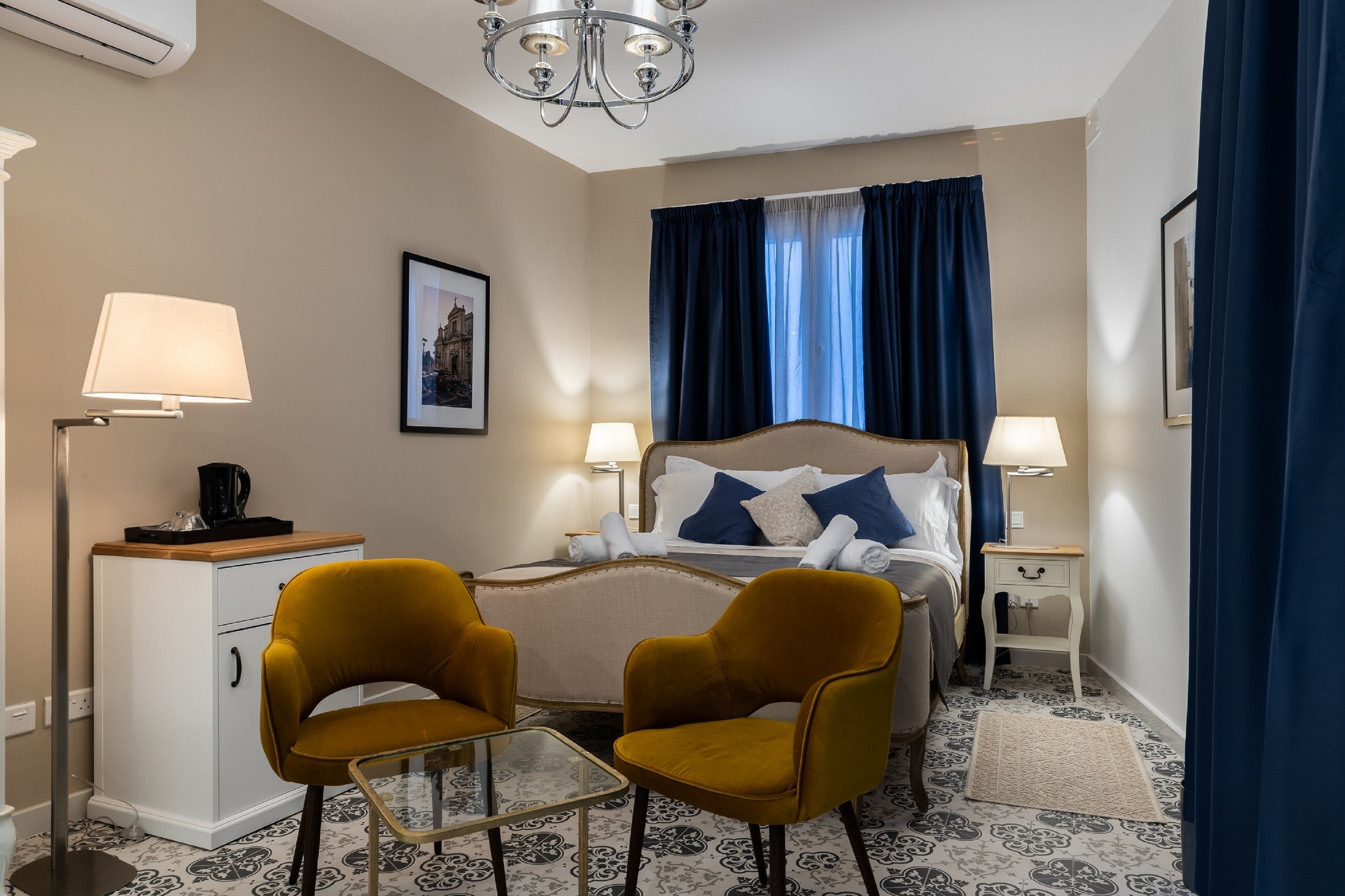 A hotel room with a bed, two chairs, a nightstand, a lamp, a picture on the wall, and a patterned rug.