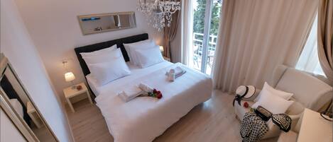 A hotel room with a large bed, a sitting area with a sofa, a chandelier, a window with curtains, and a mirror on the wall.