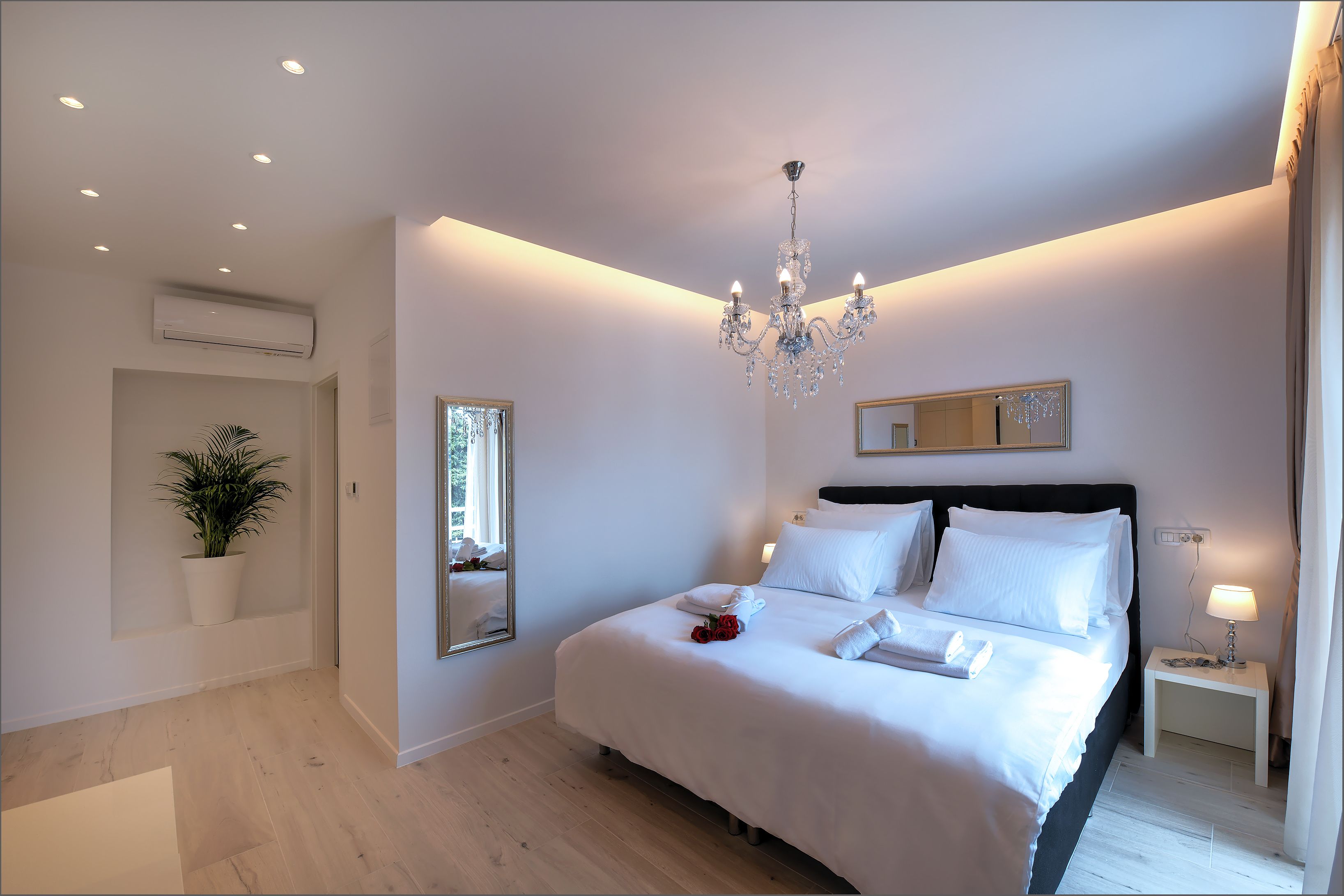 A modern bedroom with a large bed, a chandelier, a bedside table with a lamp, a potted plant, and a mirror on the wall.