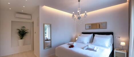 A modern bedroom with a large bed, a chandelier, a bedside table with a lamp, a potted plant, and a mirror on the wall.