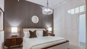 A modern bedroom with a large bed, two bedside tables with lamps, a chandelier, and a view of the outside through a window. - Clock Loft (Atlanta)