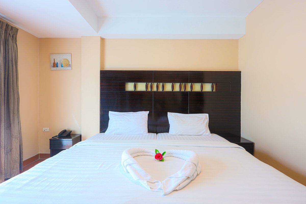 A hotel room with a neatly made bed, a heart-shaped towel arrangement, a bedside phone, and a wall-mounted light fixture.