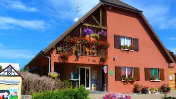Ein zweistöckiges Gebäude mit einem geneigten Dach, einem Balkon mit hängenden Blumenkörben und einem sichtbaren Eingang.