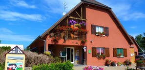 A two-story building with a sloped roof, a balcony with hanging flower baskets, and a visible entrance.