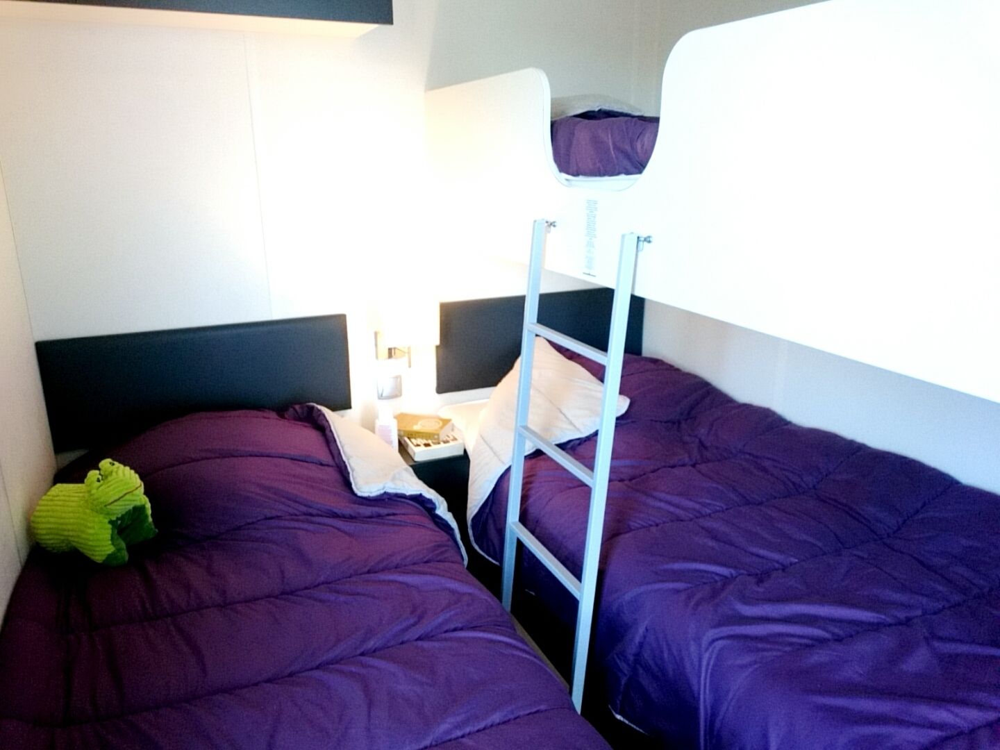 Quarto com beliche, roupa de cama roxa, escada e uma pequena mesa com um livro e uma xícara.