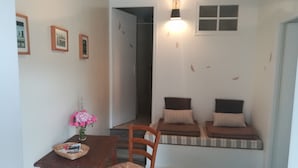Ein kleines Zimmer mit einem Holztisch, einem Korbstuhl und einer Bank mit Kissen. An der Wand hängen gerahmte Bilder und das Fenster bietet einen Ausblick.
