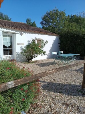 Un bâtiment blanc avec un toit en tuiles, une petite fenêtre et une porte. On y trouve une clôture en bois, un chemin de gravier et une table verte avec des chaises.