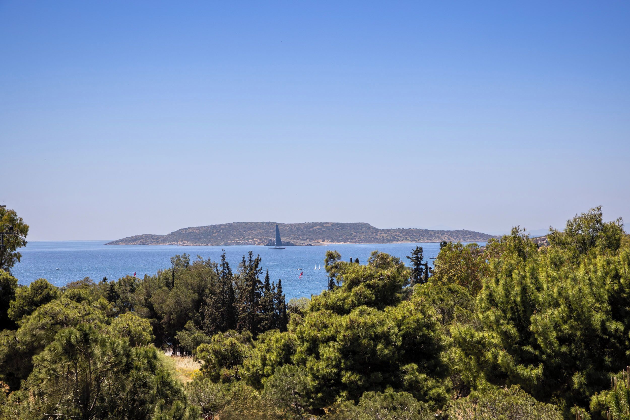 A coastal landscape with dense pine trees, a clear blue sea, and a distant island.