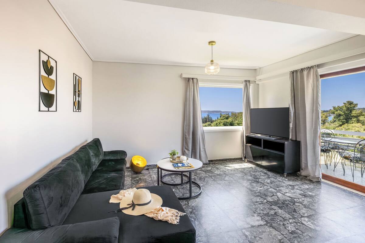 A modern living room with a black sofa, a round coffee table, and a flat-screen TV. There is a balcony with a view of the landscape.