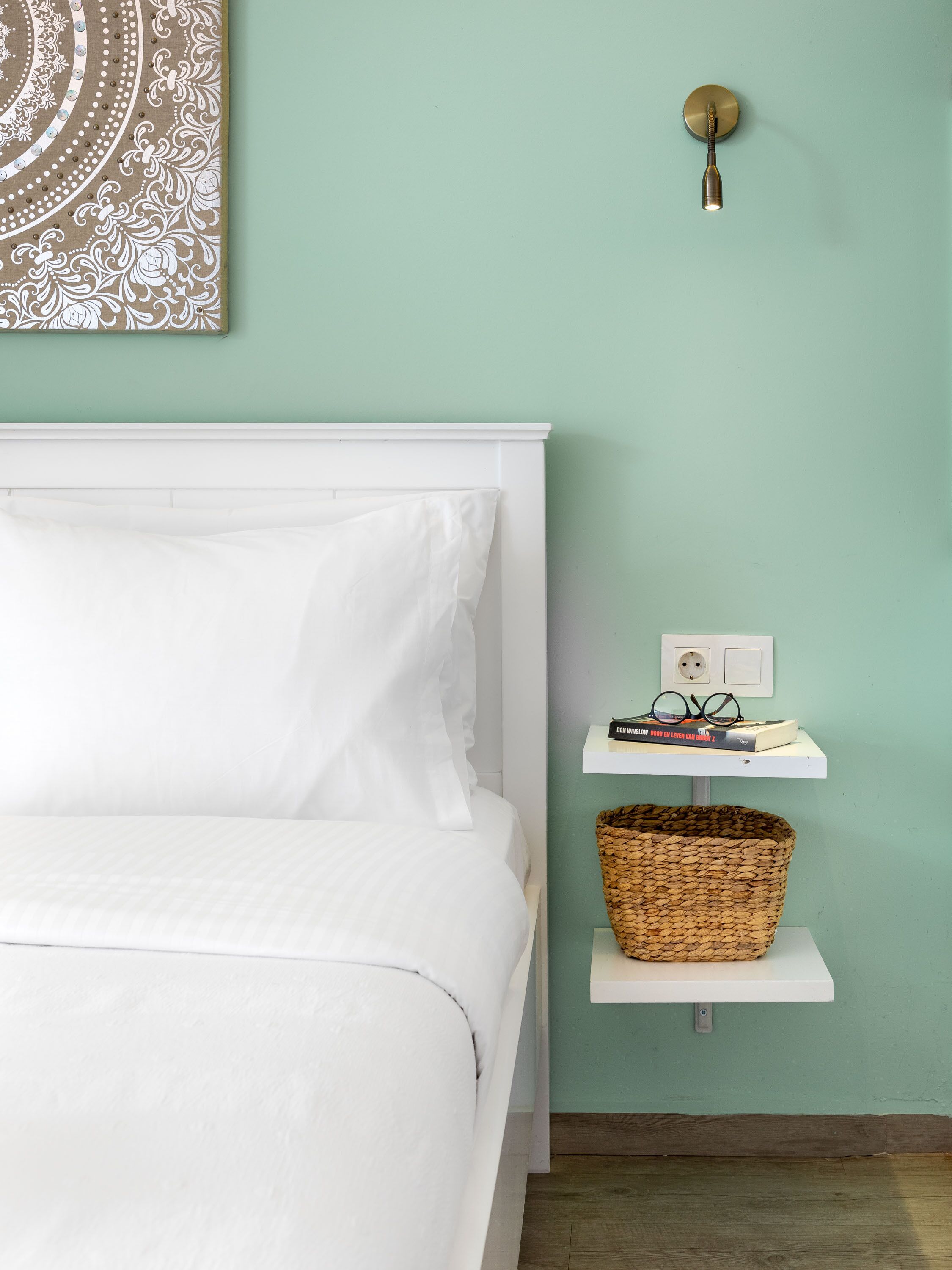 A neatly made bed with white linens, a wall-mounted light fixture, a small shelf with books and glasses, and a woven basket.
