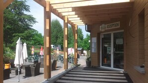 Aula, jossa on puinen pergola, ulkotila varjoisine istuimineen sekä selkeä kulkuväylä, joka johtaa sisäänkäynnille.