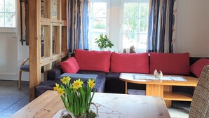 A cozy living room with a wooden ceiling, a sofa with red cushions, a wooden table with a vase of daffodils, and a window with curtains. - Ferienhof Cohrs - Ferienhäuser (Bispingen)