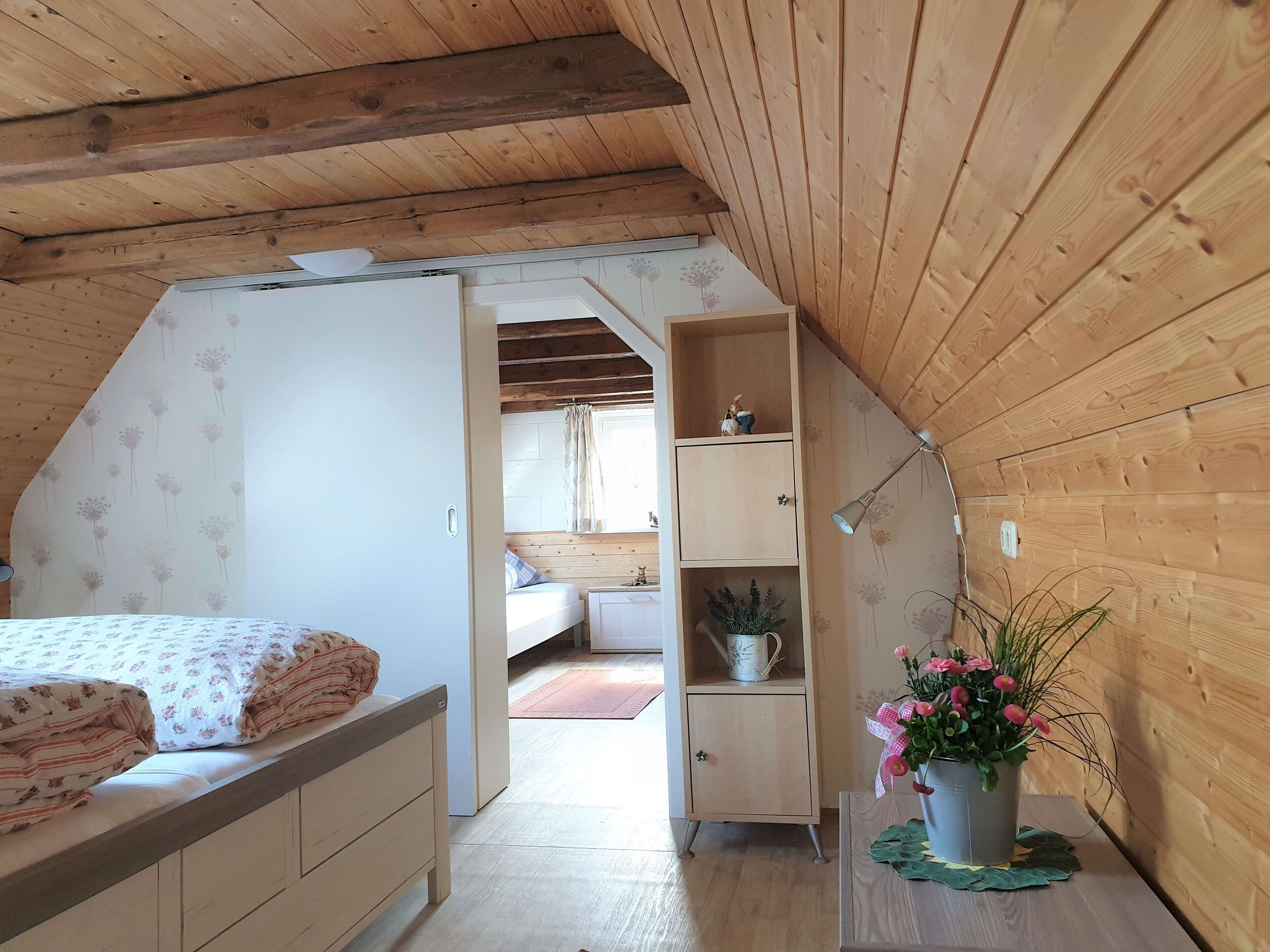 A bedroom with a sloped ceiling, wooden walls, a bed with floral bedding, a nightstand, a small cabinet, and a potted plant.