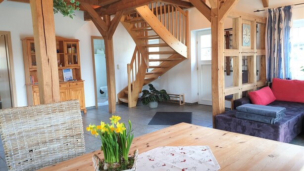 A cozy living room with a wooden staircase, a dining area with a table and chairs, and a sofa with red cushions. - Ferienhof Cohrs - Ferienhäuser (Bispingen)