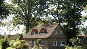 A traditional brick house with a sloped roof, multiple windows, and surrounded by lush greenery. - Ferienhof Cohrs - Ferienhäuser (Bispingen)