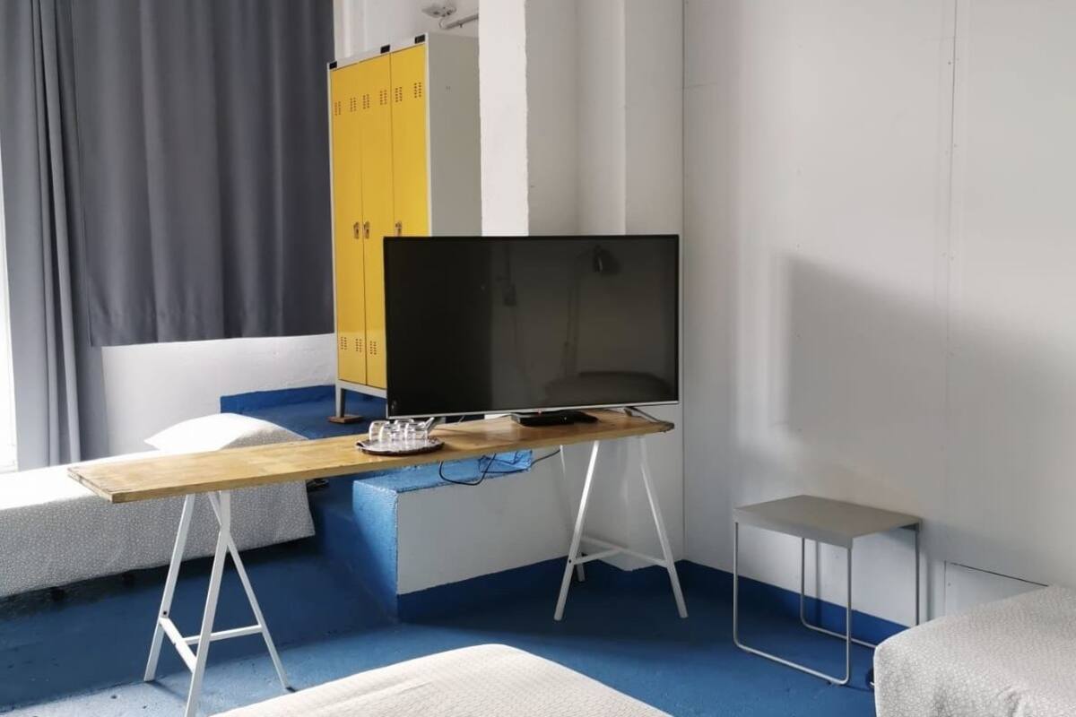 Een moderne kamer met een grote flatscreen televisie op een houten tafel, een klein bijzettafeltje, een bed met een witte matras en een blauw vloerkleed.