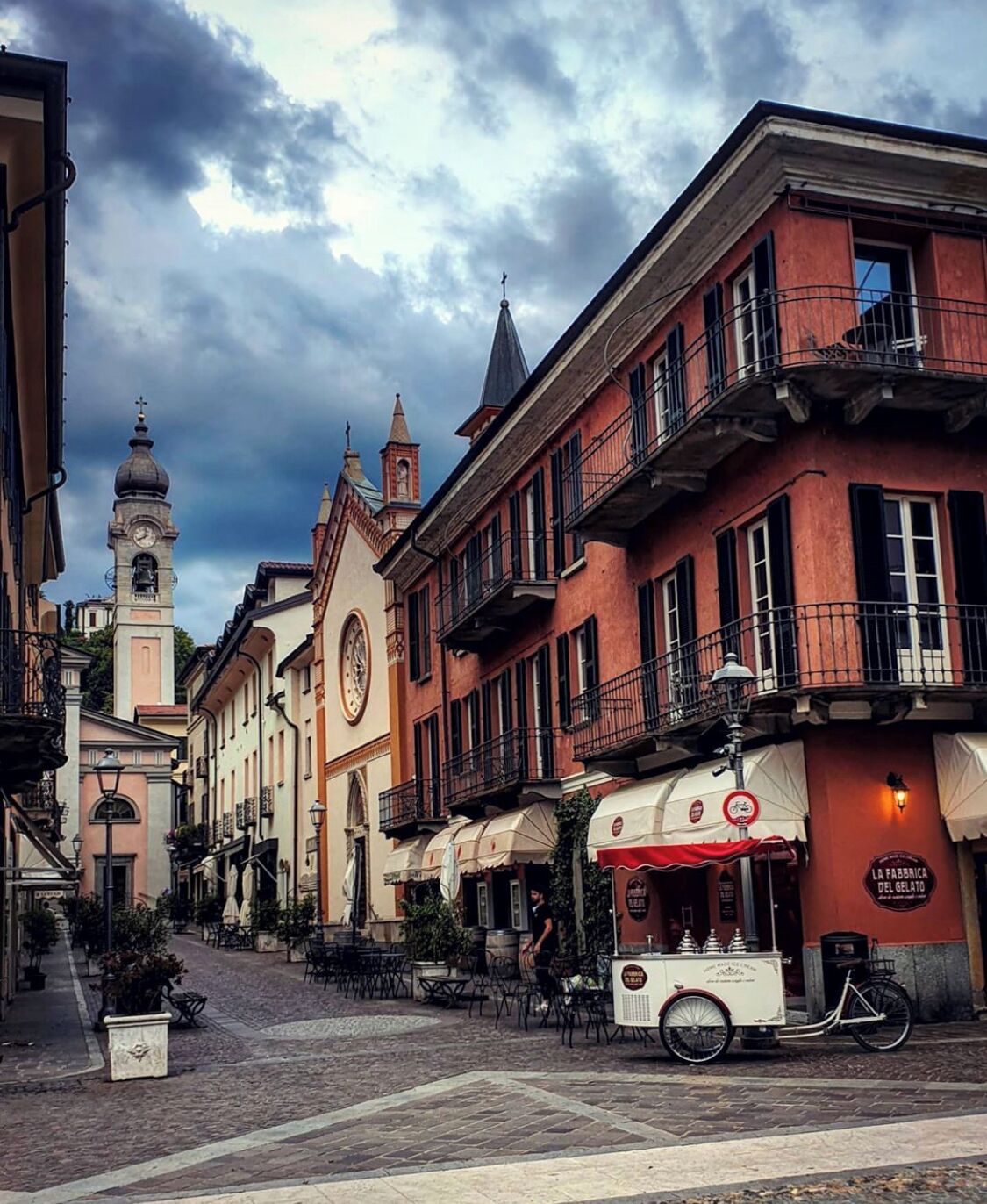 A cobblestone street lined with historic buildings, a clock tower, and outdoor seating at a gelato shop.