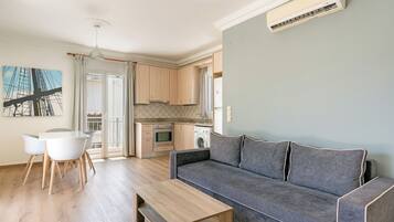 Ein modernes Wohnzimmer mit einem grauen Sofa, einem Holzesstisch und Stühlen. Im Hintergrund ist eine Küche mit Holzausbau und einer Waschmaschine zu sehen.