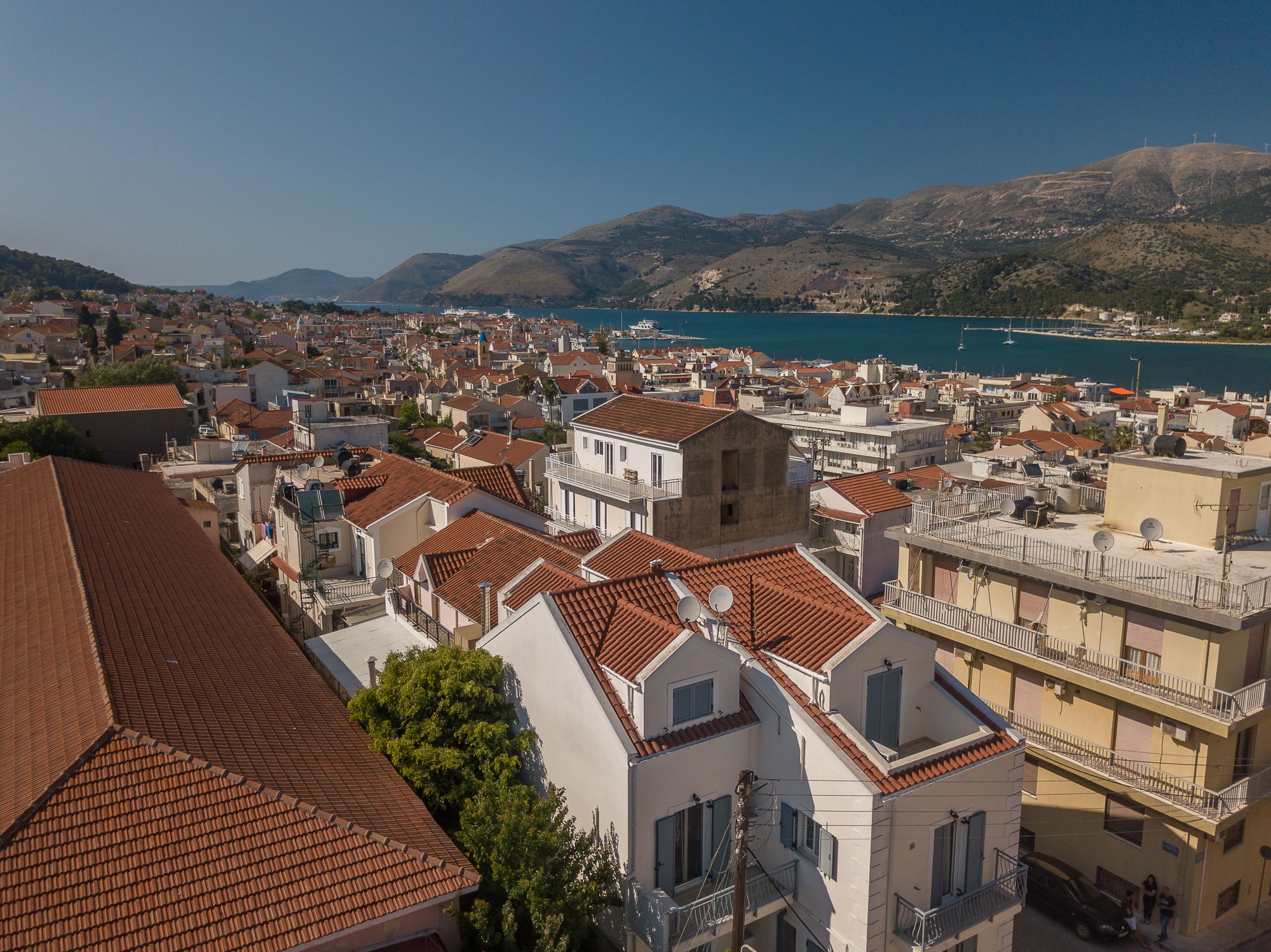A coastal town with terracotta-tiled buildings, a marina, and hills in the background.