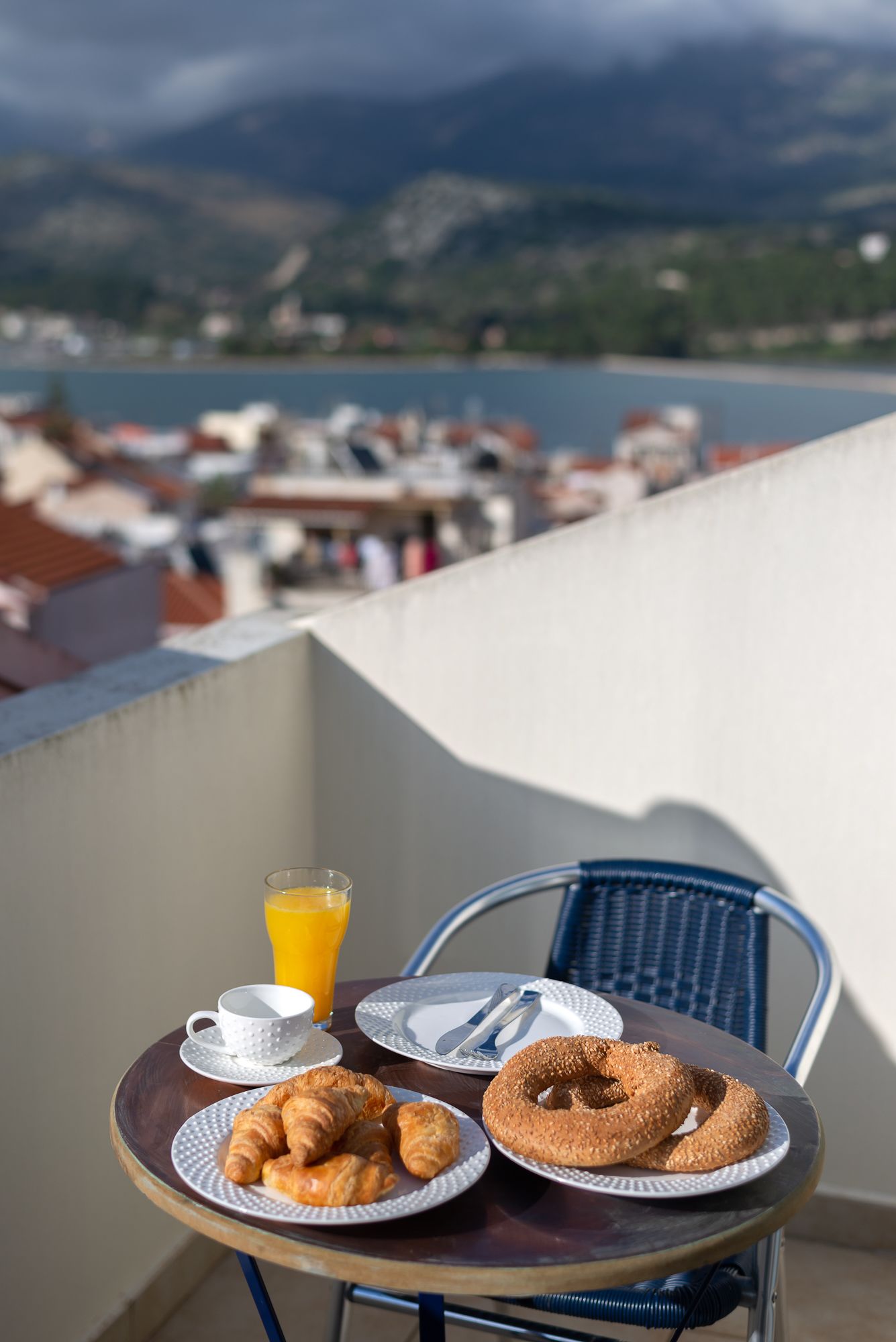 Uma mesa com croissants, um bagel, um copo de sumo de laranja e uma chávena numa varanda com vista panorâmica para um lago e montanhas.