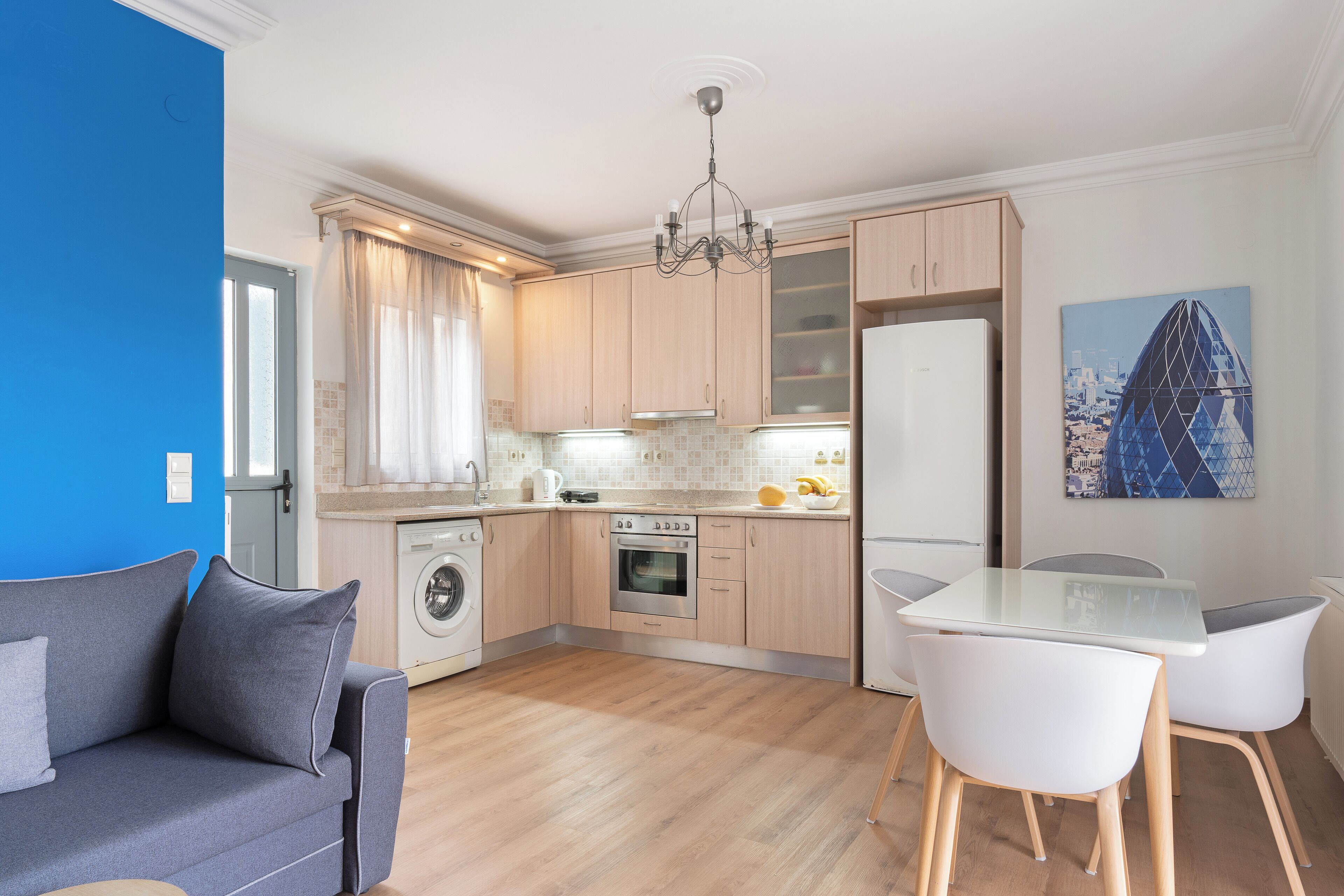 A modern kitchen with wooden cabinets, a built-in oven, and a refrigerator. There is a dining table with chairs and a sofa in the living area.