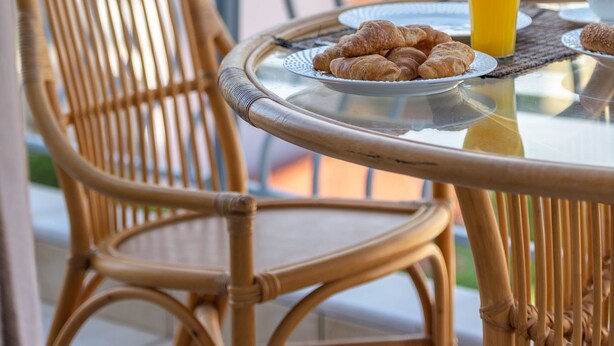 A wicker chair and a round glass table with croissants and a glass of orange juice on a balcony overlooking a coastal area.