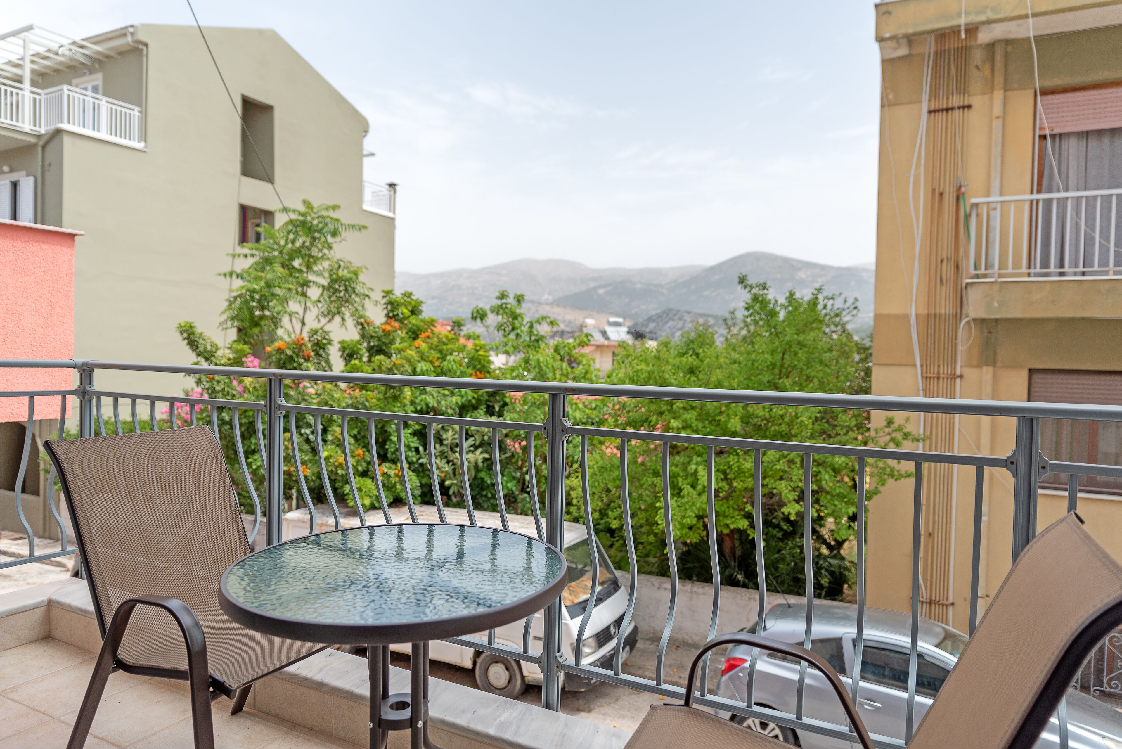 A balcony with a round table and two chairs, a parked car, and a view of distant buildings and mountains.