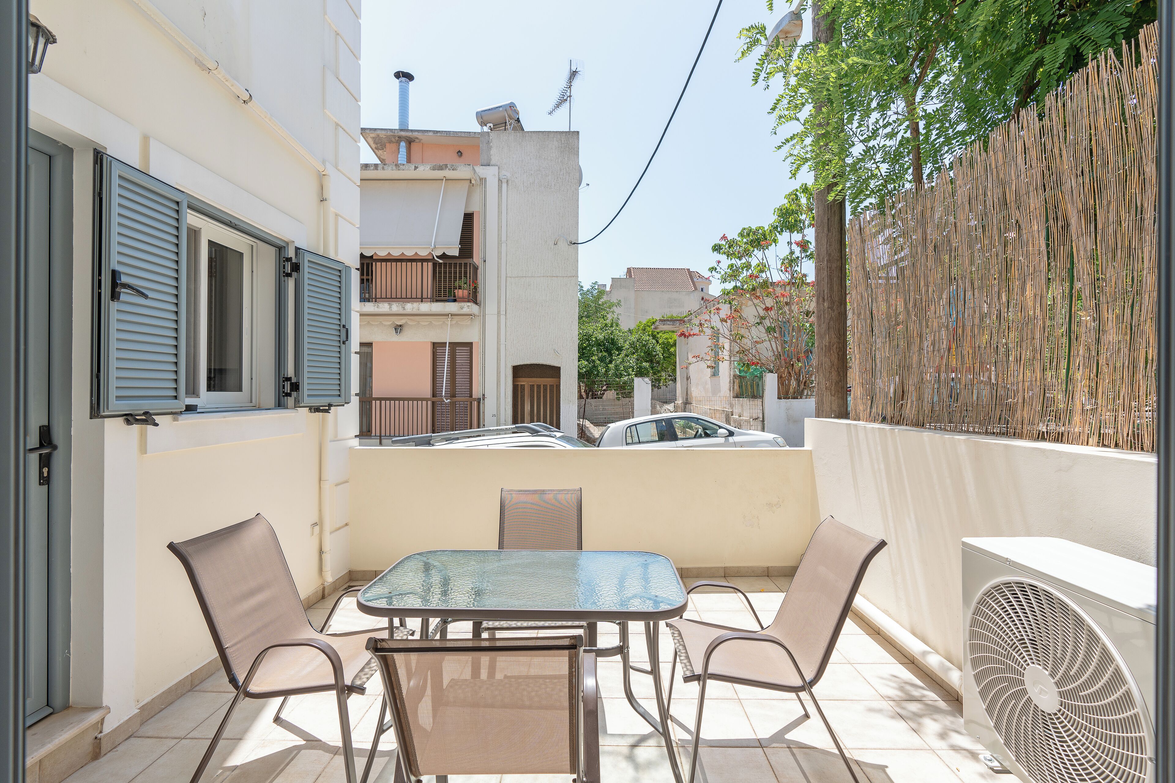 A small outdoor patio with a glass table and metal chairs, a white wall, and a bamboo fence.