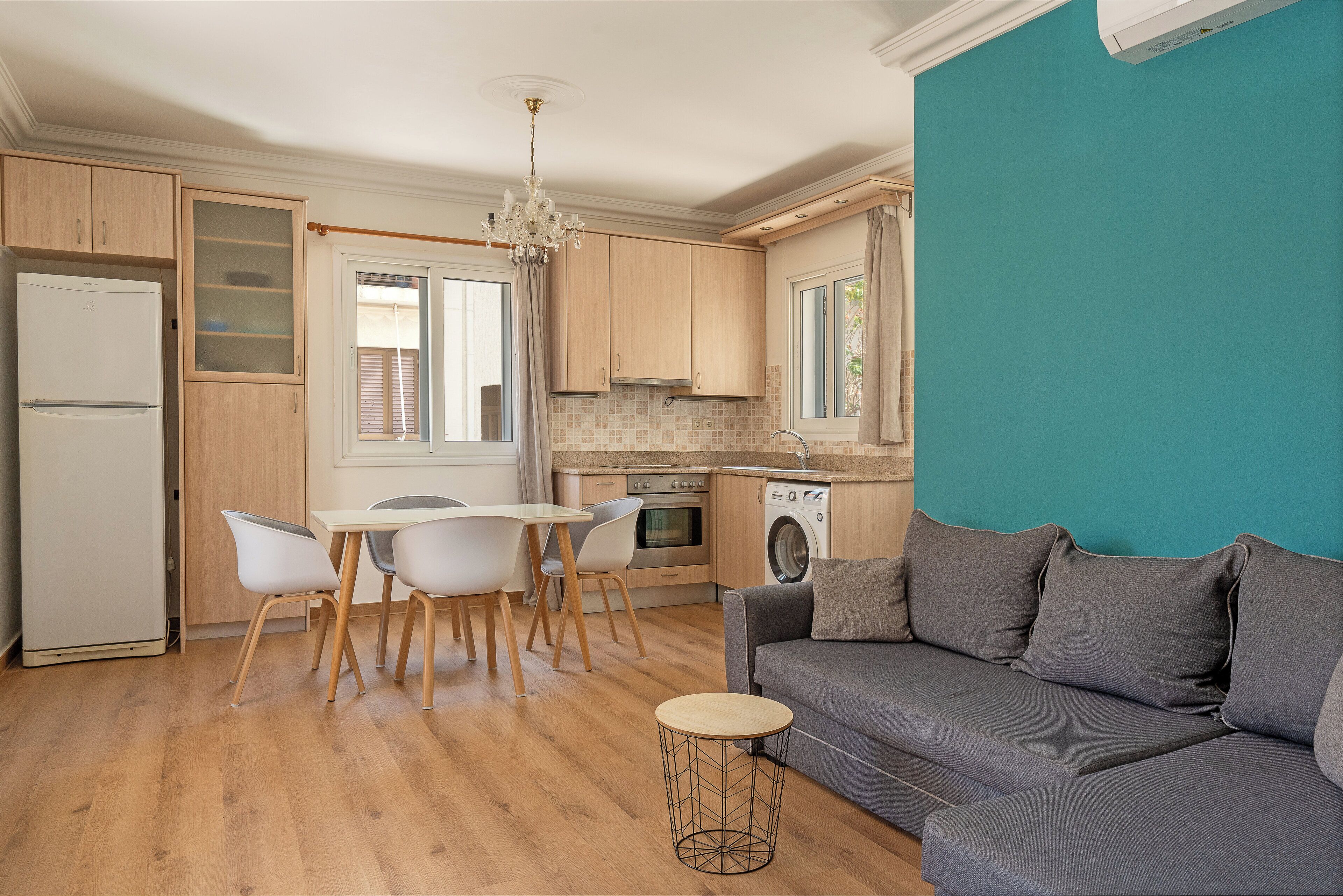 Una cocina moderna con armarios de madera, un comedor con mesa y sillas blancas, un sofá gris y una lavadora.
