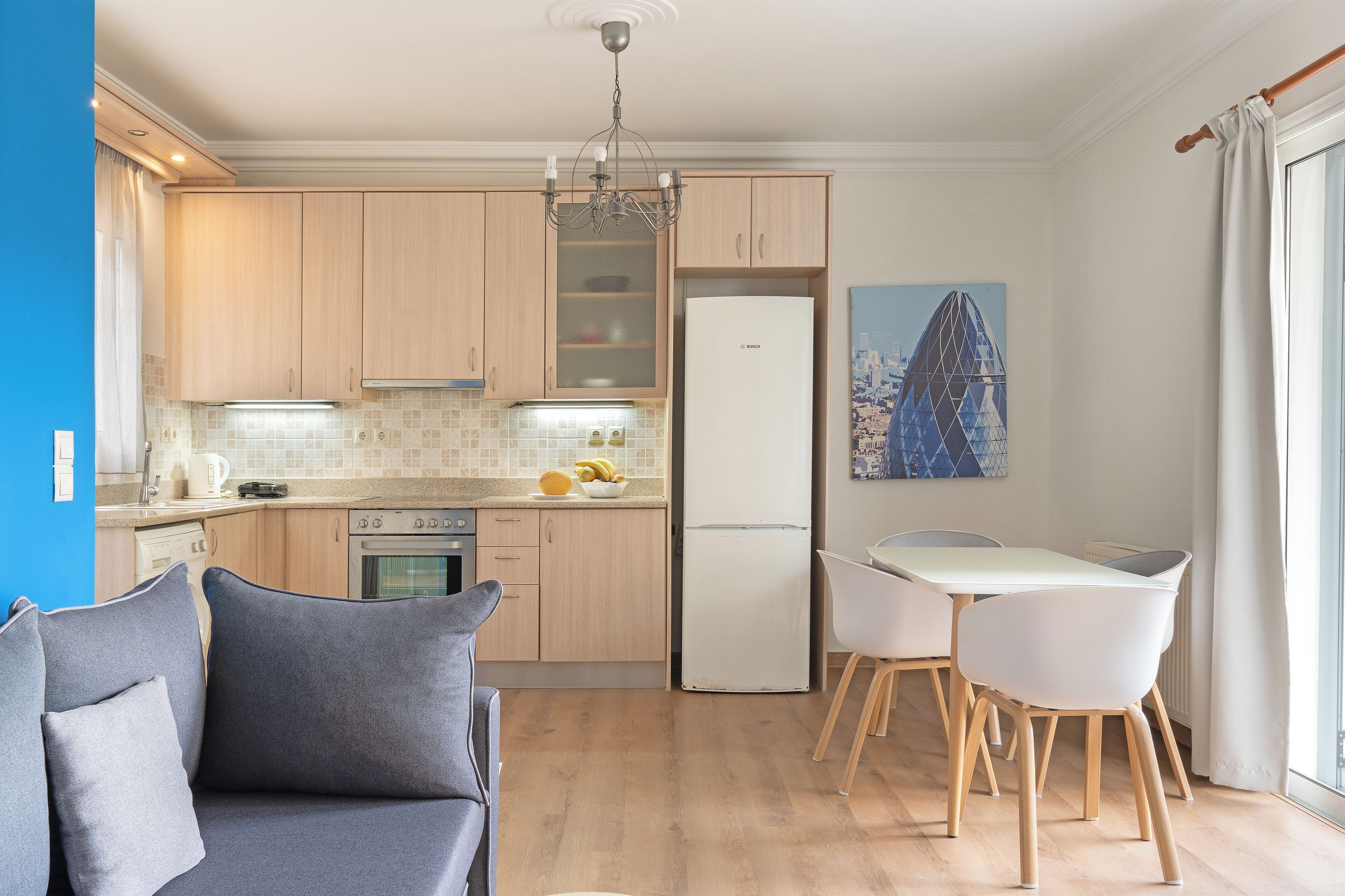 A modern kitchen with wooden cabinets, a built-in oven, and a white refrigerator. There is a dining area with a white table and chairs, and a sofa with cushions.