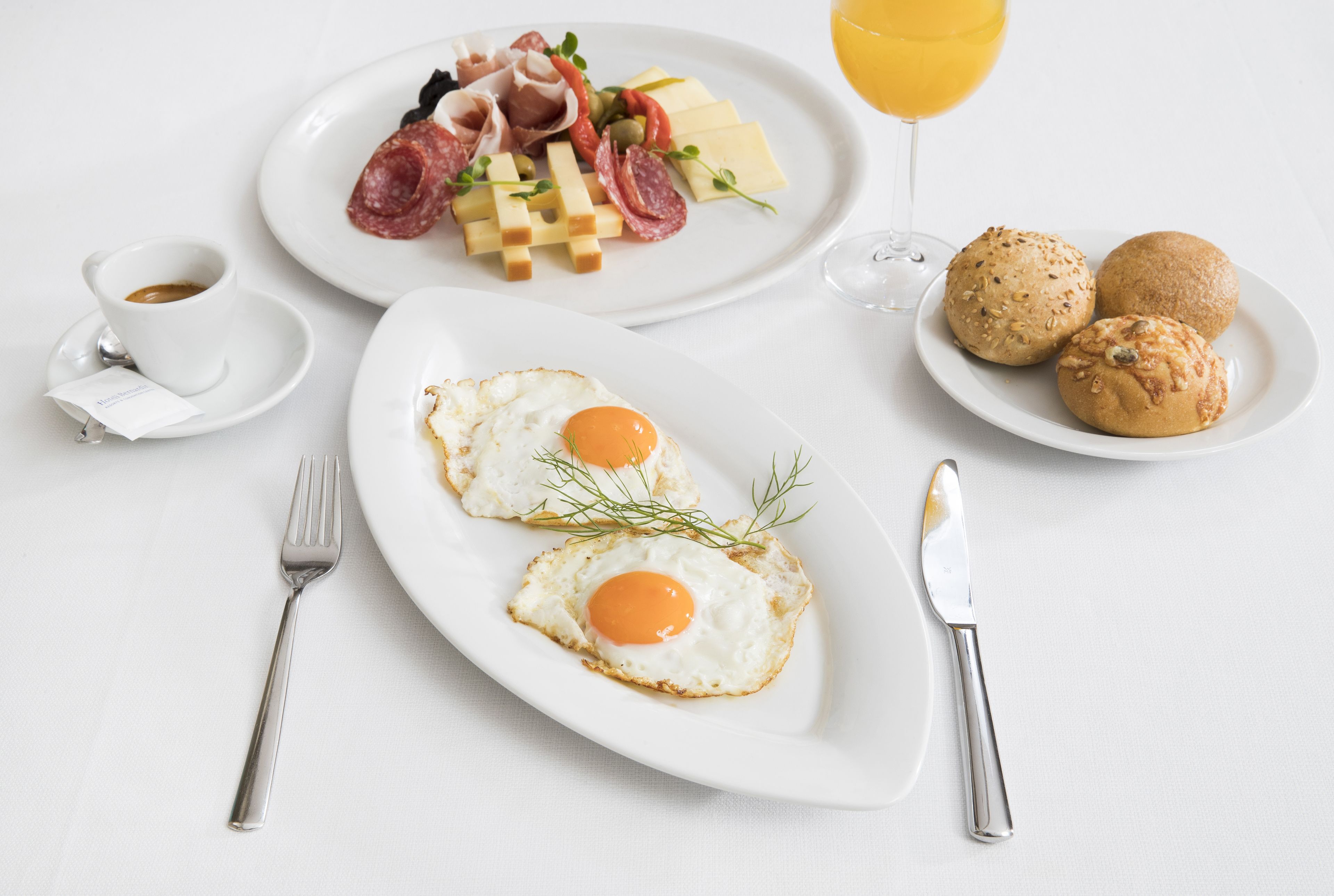 A breakfast setting with two fried eggs, a plate of meats and cheese, a glass of orange juice, and two muffins on a white table.