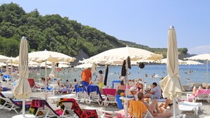 A beach with numerous sun loungers and umbrellas, some with towels on them, and a few people swimming in the water. - Hotel Haliaetum - San Simon Resort (Izola)
