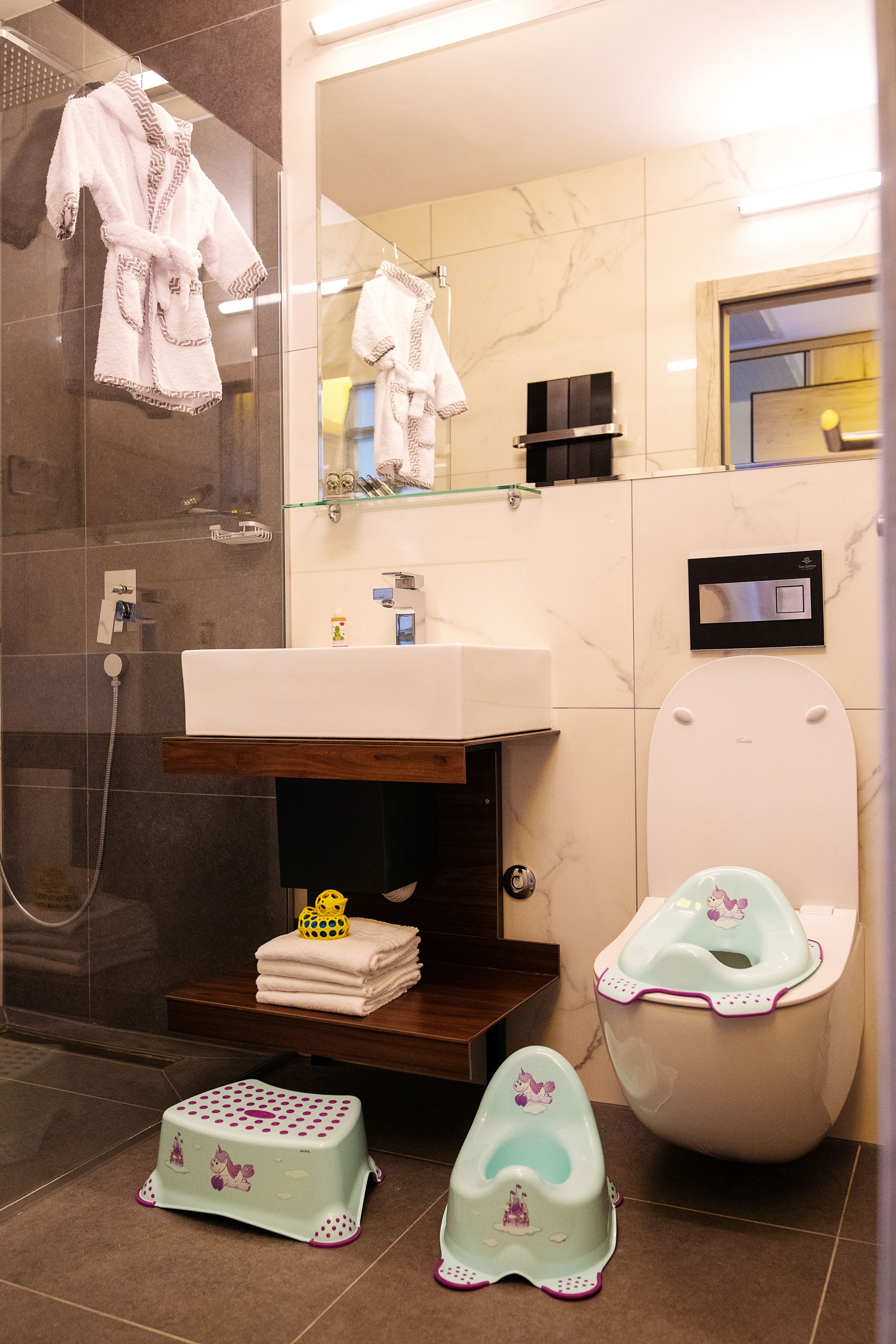 Ein Badezimmer mit einem weißen Waschbecken, einer Duschkabine aus Glas, einer Toilette mit Kindersitz und einem Spiegel, der den Raum reflektiert.