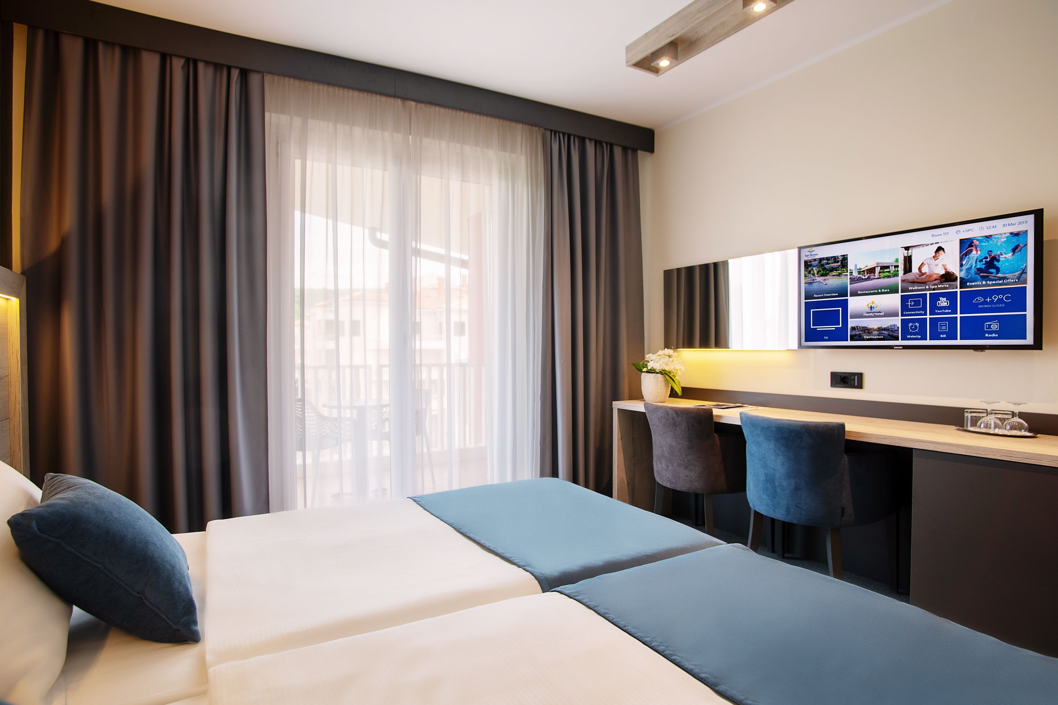Una habitación de hotel moderna con una cama, un escritorio y un televisor mostrando una aplicación del hotel.
