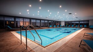 An indoor swimming pool with blue tiles, sunken seating, and a view of the city at night. - Hotel Haliaetum - San Simon Resort (Izola)