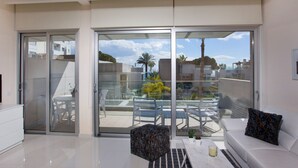 Un salon moderne avec un canapé blanc, un pouf à motifs et une porte en verre donnant sur une terrasse extérieure équipée de chaises et d’une table.