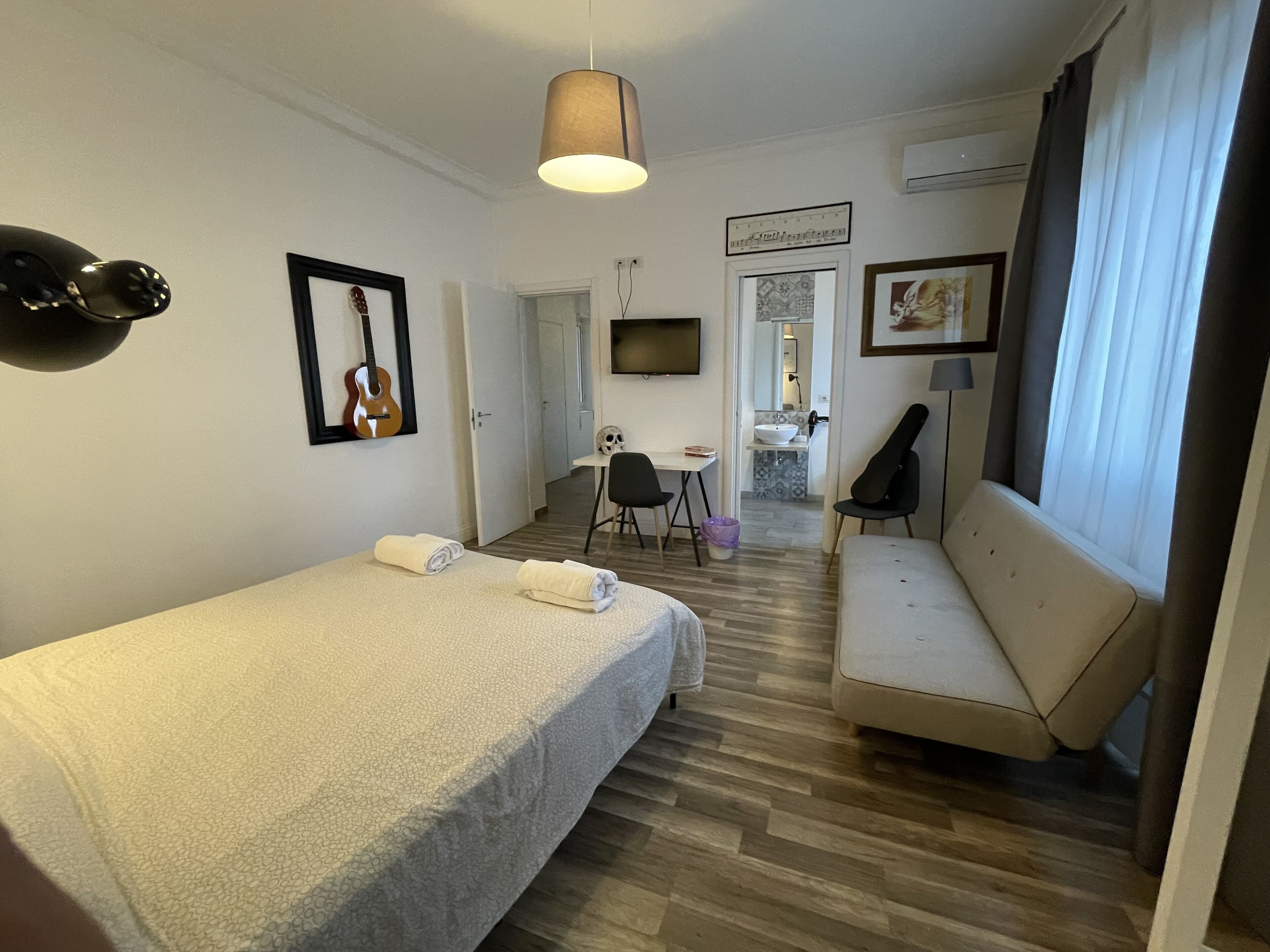A hotel room with a bed, a sofa, a desk, a chair, a TV, and a guitar on the wall.