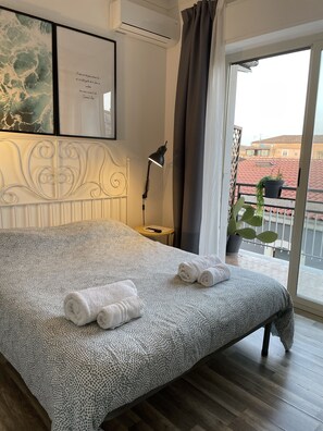 Cama arrumada com toalhas dobradas, luminária de cabeceira e vista para uma varanda com planta em vaso.