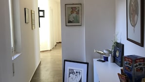 A hallway with framed artwork on the walls, a small table with a vase and a framed picture, and a box of travel brochures on a counter. - La Dimora delle Muse (Catania)