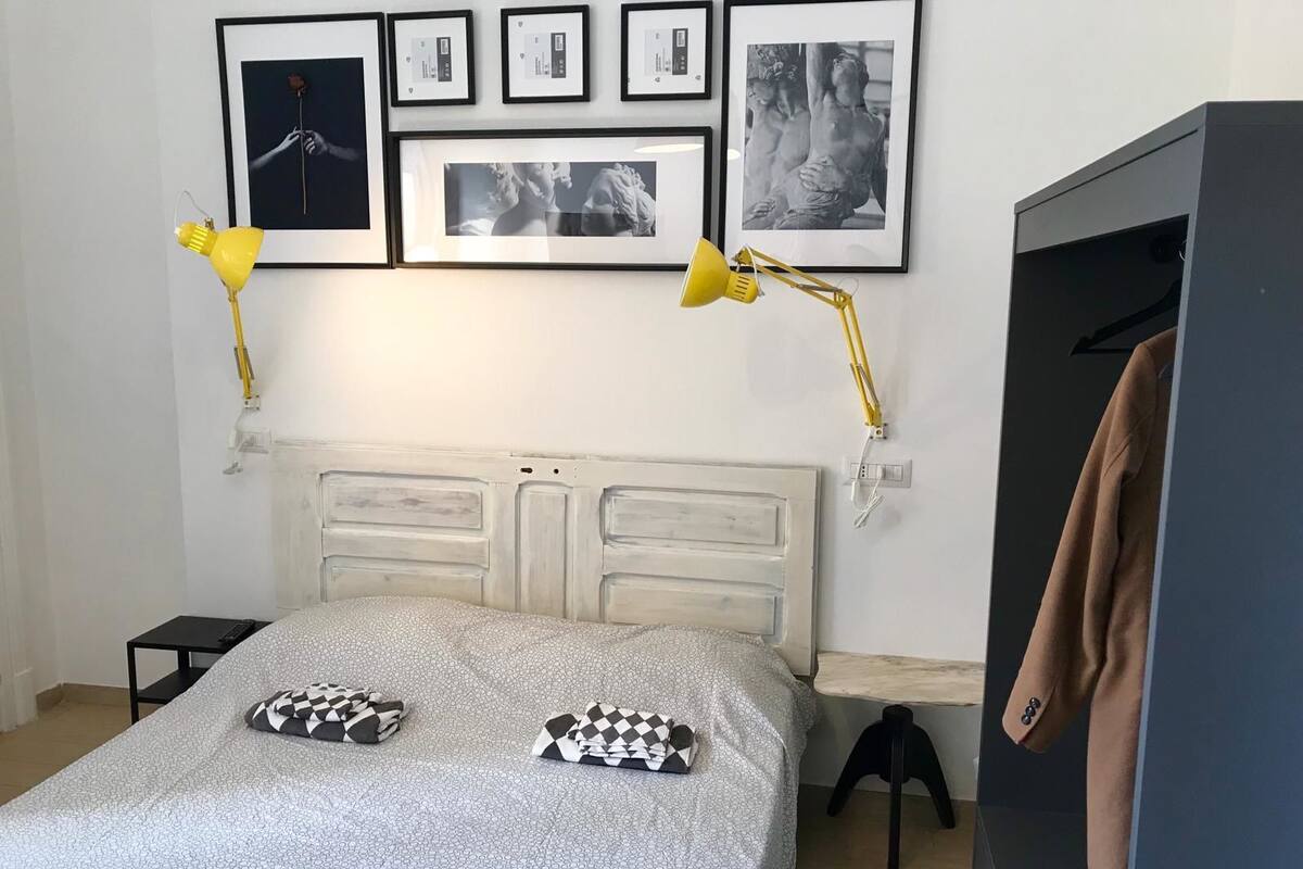 A bedroom with a bed, two pillows, a bedside table, a lamp, a wardrobe, and framed pictures on the wall.