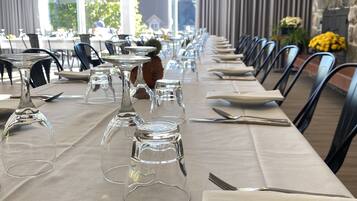 A long dining table set with clear glassware, silverware, and plates, with a stone wall and curtains in the background.