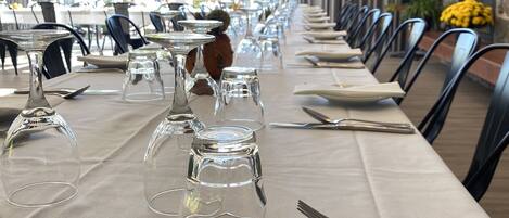 Una larga mesa de comedor dispuesta con cristalería transparente, cubiertos y platos, con una pared de piedra y cortinas al fondo.