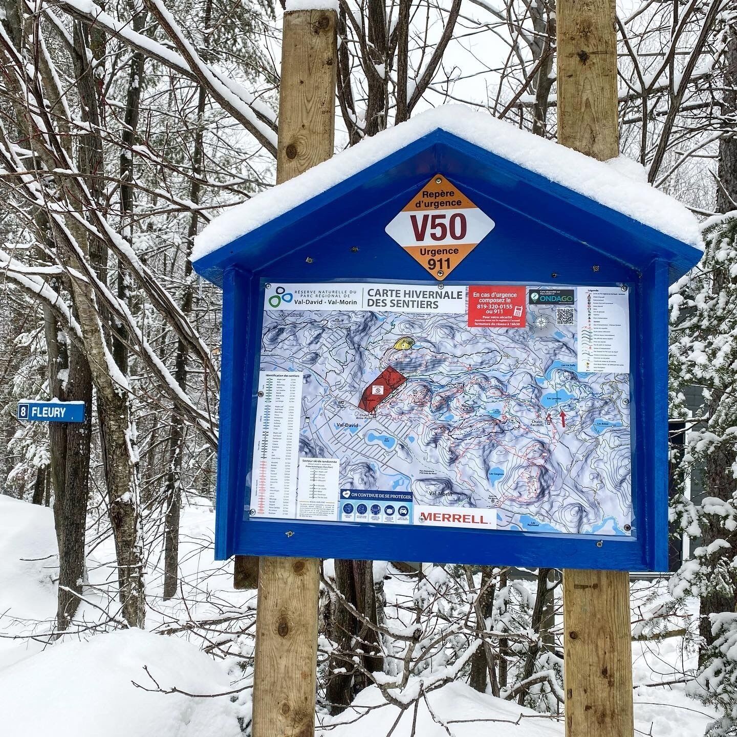 Un panneau d’information bleu, avec une carte et les coordonnées des personnes à contacter en cas d’urgence, placé dans un décor de forêt enneigée.