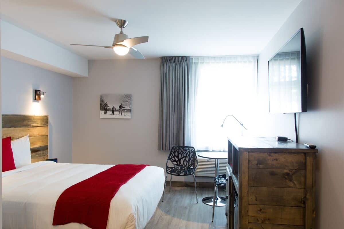 A hotel room with a bed, a desk, a chair, a ceiling fan, a TV, and a window with curtains.