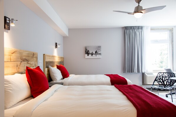 A hotel room with two beds, red and white bedding, a ceiling fan, a window with curtains, and a wall-mounted air conditioner.