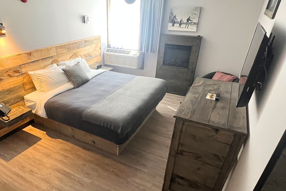 A bedroom with a wooden headboard, a bed with grey bedding, a bedside table with a phone, a wooden dresser, and a fireplace.