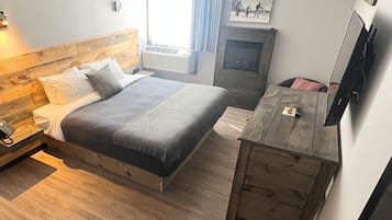 A bedroom with a wooden headboard, a bed with grey bedding, a bedside table with a phone, a wooden dresser, and a fireplace.