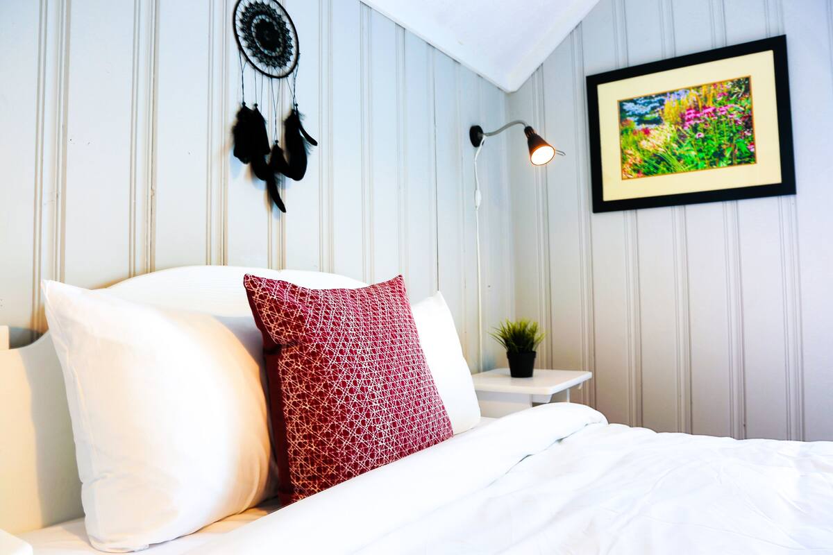 A neatly made bed with white and red pillows, a dreamcatcher, a framed picture, and a bedside lamp in a room with white paneled walls.